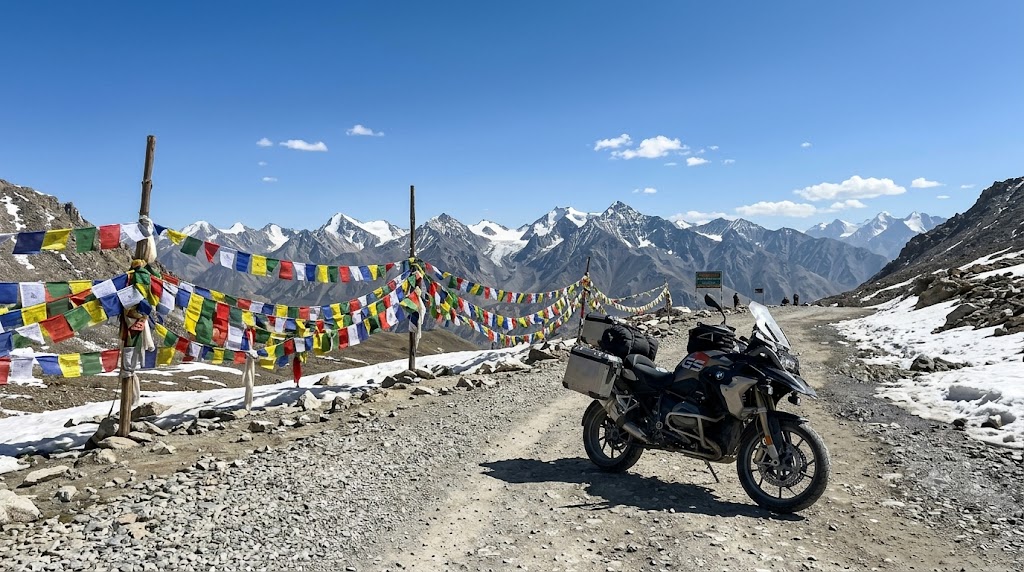 Motorcycle parked at a high mountain pass with prayer flags and snow patches near Khardung La Ladakh